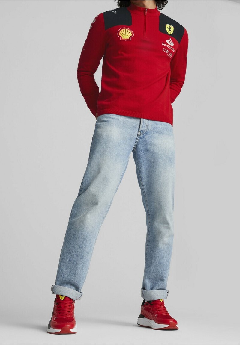 Polo rouge à manches longues avec col zippé, doté d'épaules noires et de logos, porté avec un jean en denim bleu clair et des baskets rouges.