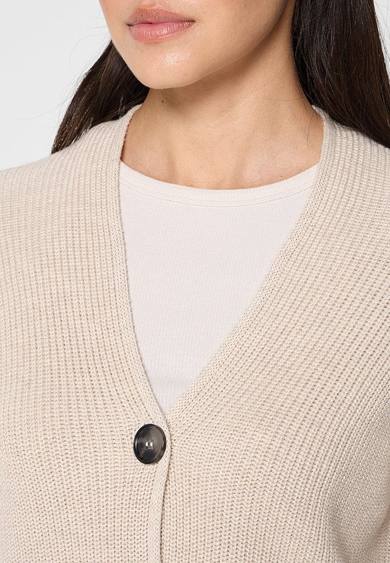 Beige gestrickte Strickjacke mit V-Ausschnitt, ausgestattet mit einem einzelnen schwarzen Knopf und einer weichen Textur. Getragen über einem hellcremefarbenen Oberteil.