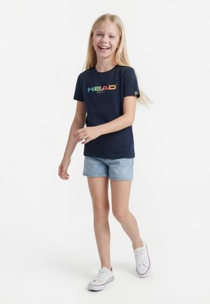 Šypsosi blondinė mergina, vilkinti tamsiai mėlyną marškinėlį su daugiapalve „HEAD“ logotipu, šviesius džinsinius šortus ir baltus sportinius batelius, žingsniuojanti pirmyn ant balto fono.