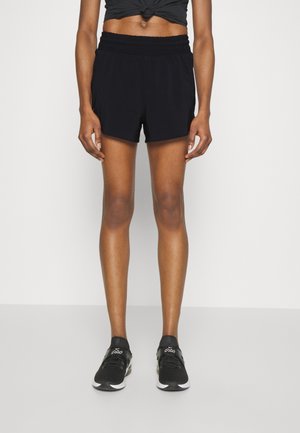 Persona que lleva pantalones cortos deportivos negros, zapatos deportivos negros y una camiseta negra corta, de pie contra un fondo liso.