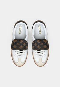 Fehér tornacipők texturált bézs orrkappal és fekete mintás pánttal. A pánton barna logók láthatók sötét háttér előtt.