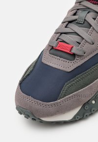 Basket en suède gris et tissu marine avec des accents verts, comportant une étiquette logo rouge, une semelle texturée et un système de laçage.