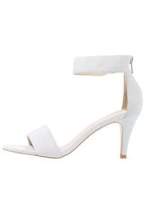 Białe zamszowe sandały damskie z odkrytymi palcami, z paskiem wokół kostki, niskim obcasem i zapięciem na suwak z tyłu.