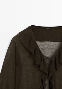 Cardigan léger, vert foncé, avec un décolleté en V et des volants le long du devant. Fabriqué en tissu texturé et transparent. S'attache au niveau du cou.