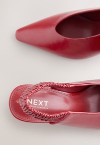 Chaussure à talon haut en cuir rouge avec un bout pointu, des côtés ouverts et des accents froncés sur le talon. La semelle intérieure porte la marque "NEXT Forever Comfort".