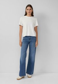 Biała koszulka z krótkim rękawem o luźnym kroju, zestawiona z jasnoniebieskimi dżinsami o szerokich nogawkach oraz białymi trampkami z beżową podeszwą, stojąca na neutralnym tle.