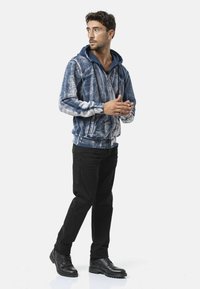 Capuchonvest met ritssluiting in blauw-wit abstract patroon, fleece materiaal, gecombineerd met zwarte broek en zwarte laarzen. Heeft een losse pasvorm.