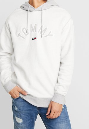 Person iført en hvid Tommy-hoodie med grå manchetter og kant, og slidte blå jeans, med den ene hånd i lommen.