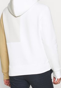 Persona che indossa un hoodie a blocchi di colore con sezioni bianche, beige e marrone chiaro, abbinato a pantaloni a quadretti scuri, visto da dietro.