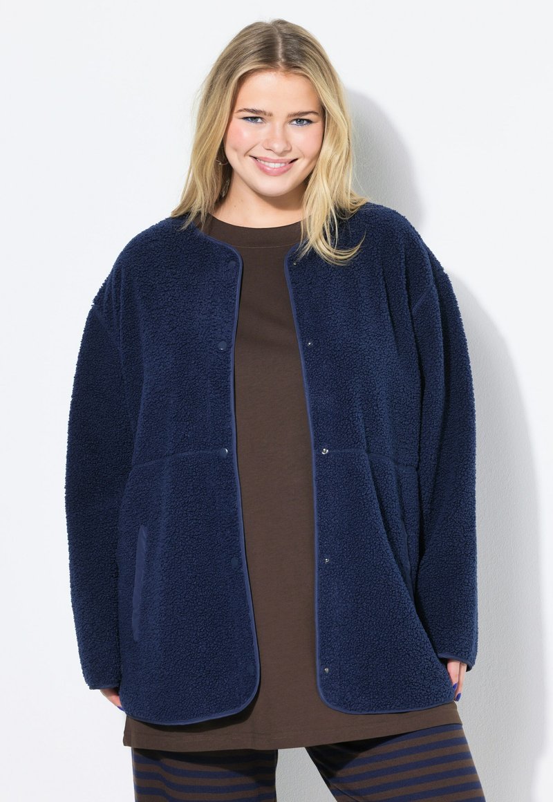 Lächelnde Frau, die eine marineblaue Fleecejacke über einem braunen Oberteil und einer marinen und braunen gestreiften Hose trägt, steht vor einem weißen Hintergrund.