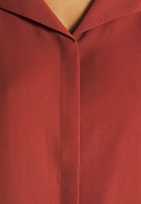 Blouse bordeaux avec une texture lisse, dotée d'un col pointu et d'un système de fermeture à boutons à l'avant. Le tissu semble léger et fluide.