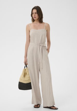 Žena stojaca v béžovom ľanovom bezrukávovom overal s opaskom v páse, drží čierno-béžovú pletenú tašku, má na sebe čierne sandále.