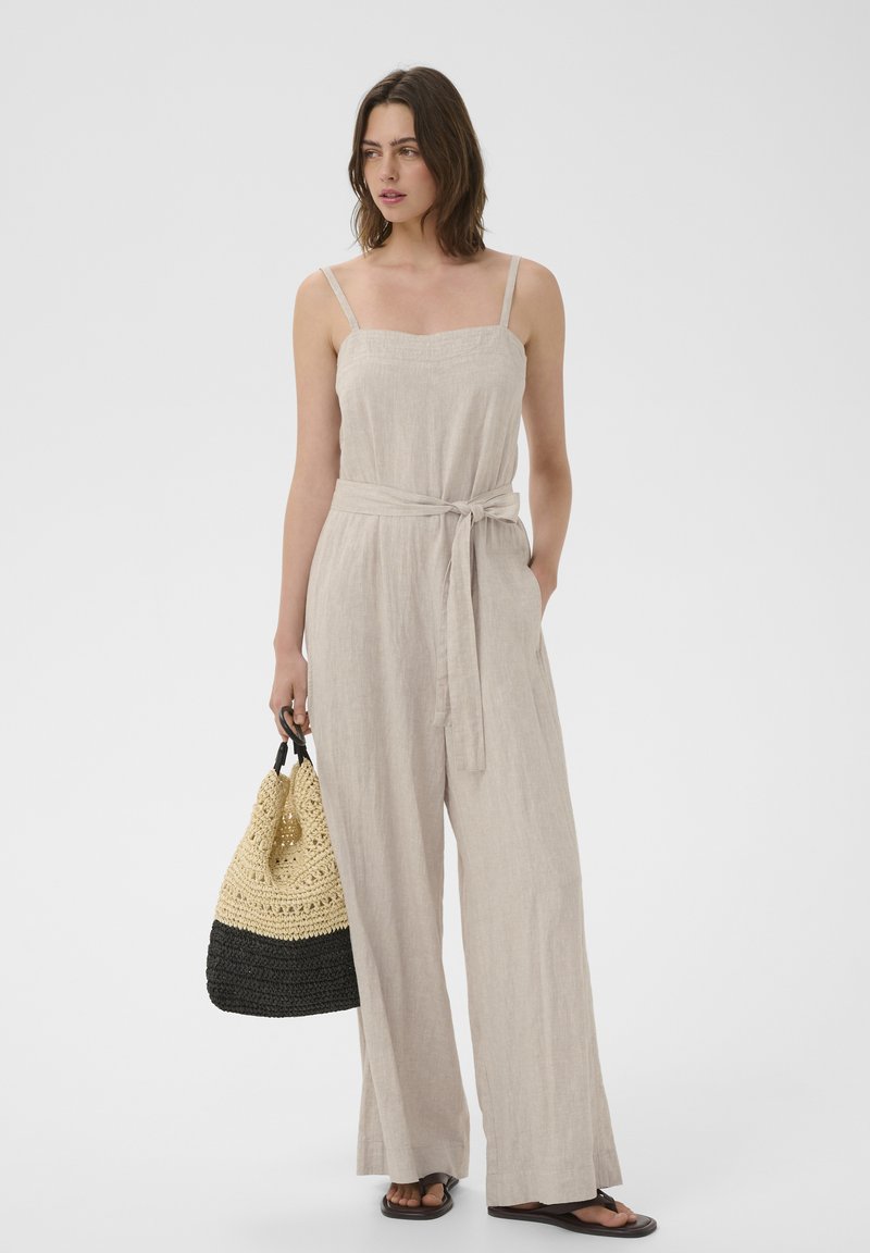 Femme debout portant une combinaison sans manches en lin beige avec ceinture à nouer, tenant un sac tissé noir et beige, portant des sandales noires.