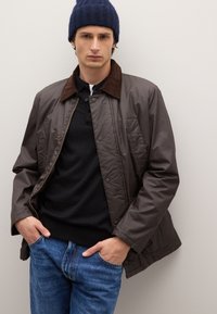 Giacca marrone leggera con colletto in velluto a coste, bottoni a pressione e due tasche laterali, indossata sopra un maglione nero e jeans blu.