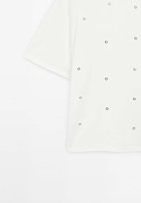 Chemise blanche à manches courtes avec de petits œillets métalliques argentés éparpillés de manière aléatoire sur le tissu.