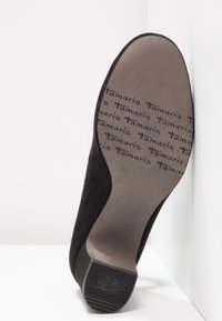 Semelle d'une chaussure à talon haut noire montrant le marquage "Tamaris" répété et un bout de talon antidérapant avec des rainures texturées.