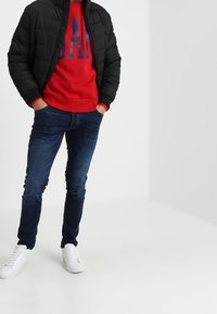 Zwart doorgestikt jack, rode sweatshirt met "GAP"-logo, donkerblauwe jeans, witte sneakers. Casual outfit met contrasterende kleuren en texturen.