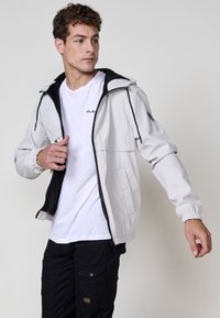 Giacca a vento grigio chiaro con interno nero, tasche frontali, polsini elastici e chiusura con zip. Indossata sopra una maglietta bianca.