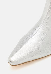 Botte argentée à bout pointu avec une finition métallique craquelée. Présente une texture lisse et une silhouette élancée. Talon empilé non visible.