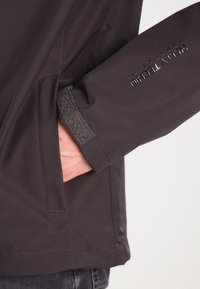 Black waterproof jacket with a textured sleeve cuff, featuring a side pocket and logo detail on the arm. Smooth exterior finish.