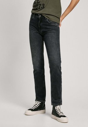 Persoon die donkere slim-fit jeans draagt, zwarte hoge sneakers met witte veters en zolen, en een olijfgroen shirt.