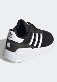 Svart atletisk sko med överdel i mesh, vit tre-stripesdesign, texturerade svarta detaljer och en vit gummisula.