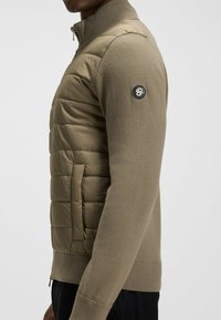 Hybrid men's jacket in khaki, featuring a quilted padded section and knitted sleeves. Zipper closure and logo patch on the left sleeve.