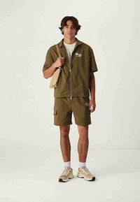 Ελιόχρωμο φερμουάρ κοντομάνικο μπουφάν και ταιριαστά σορτς, και τα δύο με λειτουργικές τσέπες. Φοριέται με μπεζ αθλητικά παπούτσια και καφέ τσάντα ώμου.