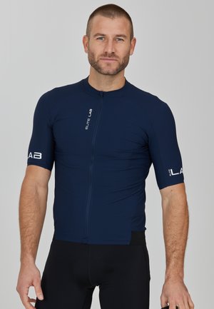 Maillot de cyclisme bleu marine à manches courtes, texture lisse, fermeture éclair à l'avant et accents de logo sur les manches. Porté avec un short de cyclisme noir.
