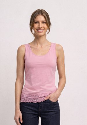 Femme souriante portant un haut sans manches rose avec un bord en dentelle et un jean foncé, debout avec une main dans la poche devant un fond uni.