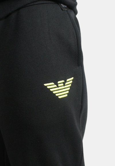 Joggers neri in morbido tessuto con un logo a strisce gialle ben visibile sulla coscia sinistra e tasche laterali per la funzionalità.