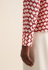 Camicia a maniche lunghe con motivo ondulato rosso e marrone scuro su tessuto bianco. Presenta polsini con bottoni e una texture liscia.