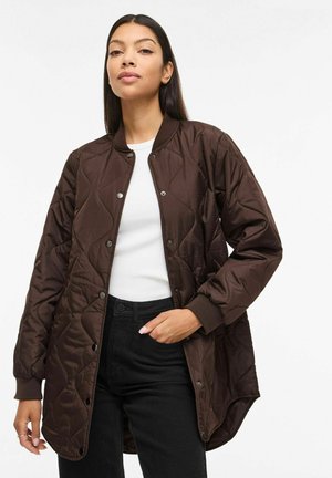 Vrouw draagt een lange donkerbruine doorgestikte jas met drukknopen, een wit topje en een zwarte broek, staand tegen een witte achtergrond.