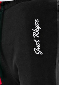 Black joggers made of soft fabric, featuring a white "Just Rhyse" logo and contrast panel along the side. Simple design with elastic waistband.