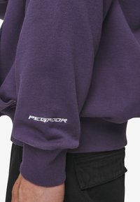 Hanorac violet din bumbac, cu manșete ribbate și un logo alb pe mânecă. Materialul pare moale, cu o textură netedă.