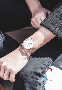 Reloj de malla en oro rosa con una esfera blanca y marcadores de hora en plata, combinado con una pulsera metálica simple en una muñeca de tono de piel claro.