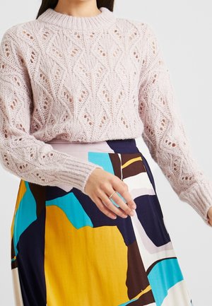 Pull en maille rose pâle avec un motif en diamant, col et poignets côtelés. Associé à une jupe colorée à motif avec des couleurs bleue, jaune et marron.