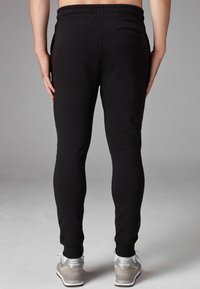 Schwarze Jogginghosen mit schlanker Passform, elastischem Bund und elastischen Bündchen. Mit einer Gesäßtasche und glattem, strukturiertem Stoff. Dazu graue Sneaker.