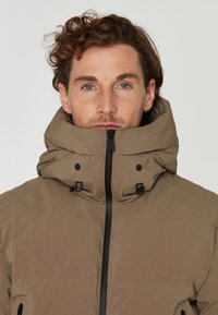 Brun, isolerad jacka med hög krage, svarta dragkedjor, sidofickor och justerbara detaljer. Slät, matt yta och minimalistisk design.
