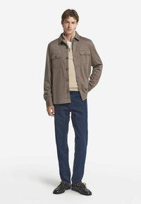 Brun knapp-up skjorta med två bröstfickor, kombinerad med en beige topp och mörka jeans, avslutad med svarta snörskor.