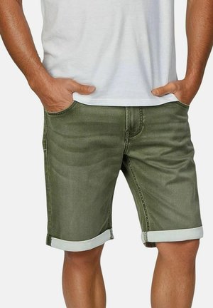 Uomo che indossa pantaloncini di jeans verde oliva risvoltati e una maglietta bianca, in piedi con le mani nelle tasche.