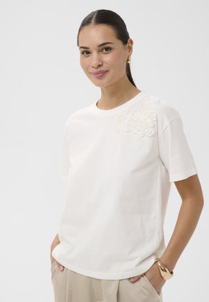 Donna con capelli scuri raccolti in una coda di cavallo, che indossa una maglietta bianca a maniche corte con applicazioni floreali sulla spalla, pantaloni beige e un braccialetto d'oro.