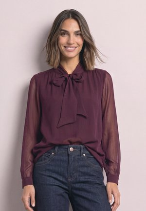 Femme souriante portant un chemisier violet transparent à manches longues avec un nœud au cou et un jean bleu foncé taille haute.