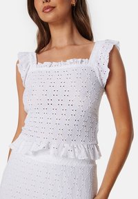 Bílý top s výstřihem do čtverce, s volánovými ramínky, texturovanou látkou a frill lemem. Vzor zahrnuje detaily s výřezy a vrstvený design.