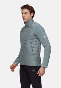 Veste isolante gris clair avec un col montant, dotée de panneaux avant matelassés et de manches en tissu doux. Logo sur la poitrine gauche et la manche.