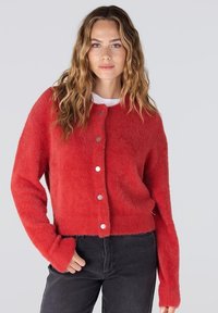 Rode pluizige cardigan met een ronde hals, zilveren knopen aan de voorkant en lange mouwen. Draagt over een wit topje en gecombineerd met donkere spijkerbroek.