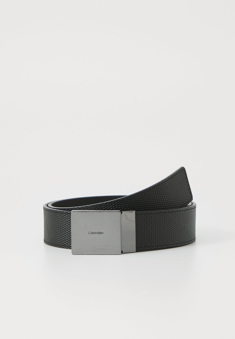 Zwarte gestructureerde riem van synthetisch materiaal met een rechthoekige zilveren metalen gesp waarop de merknaam "Calvin Klein" staat.