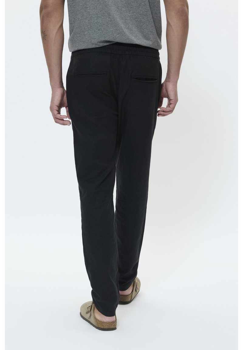 Homme portant un pantalon noir, un t-shirt gris et des chaussures beige sans lacets, debout, dos tourné sur fond blanc.