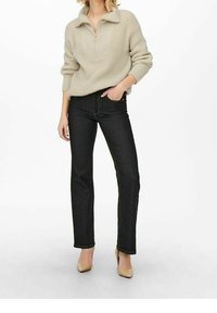 Pull en tricot beige avec fermeture éclair à l'avant, associé à un jean en denim noir et des escarpins nude. La tenue présente une coupe décontractée et un tissu texturé.