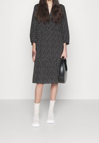 Schwarzes Kleid mit einem geometrischen weißen Muster, elbengraue Ärmel und einem gestuften Rock. Kombiniert mit weißen Stiefeln in Knöchelhöhe und einer schwarzen Tasche.